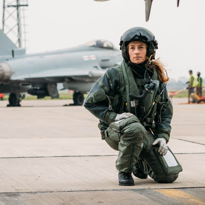 Female RAF fighter pilot kneeing on concrete apron inspecting underneath of aircraft before flight with Typhoon in background