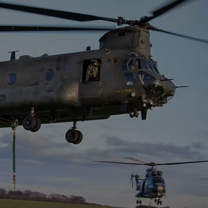 RAF Weapon systems operator leaning out of RAF Chinook side door, monitoring load-slinging, while exercising with RAF Puma